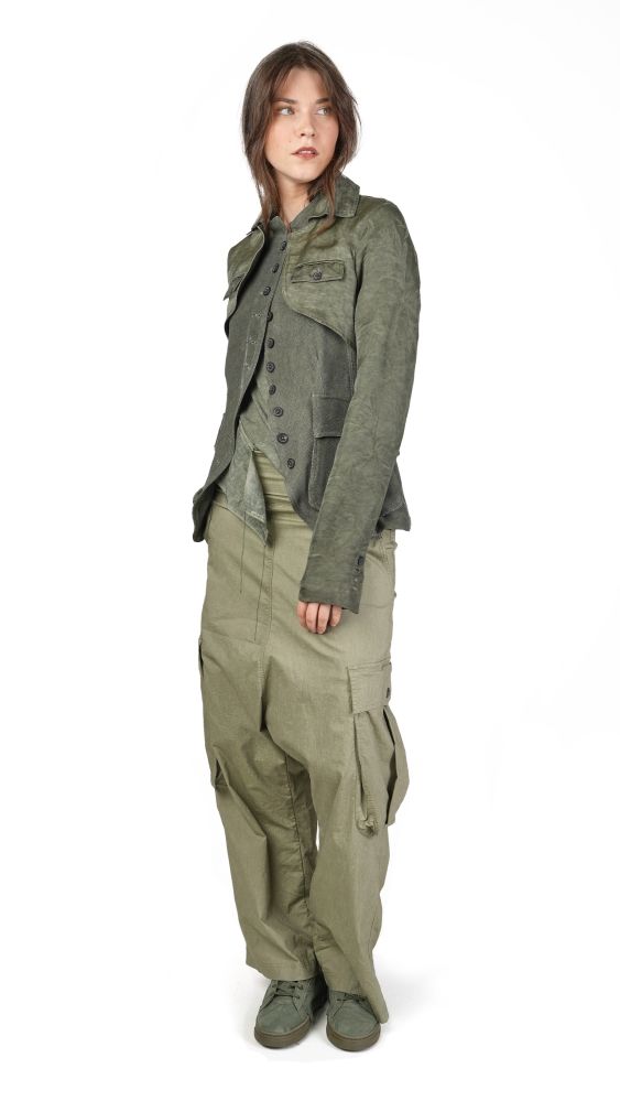 Person trägt olivgrüne Arbeitsjacke mit weiten Hosen und festem Schuhwerk, seriös-distanzierter Blick.