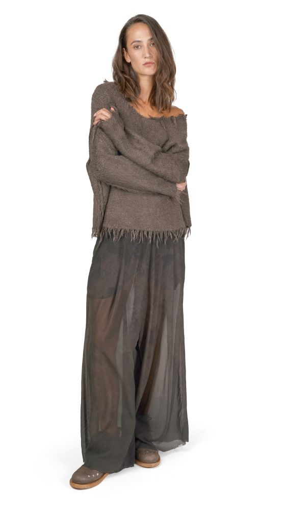 Frau trägt oversized grauen Pullover mit Fransen, schwarze weite Hosen und flache Schuhe, Arme verschränkt.